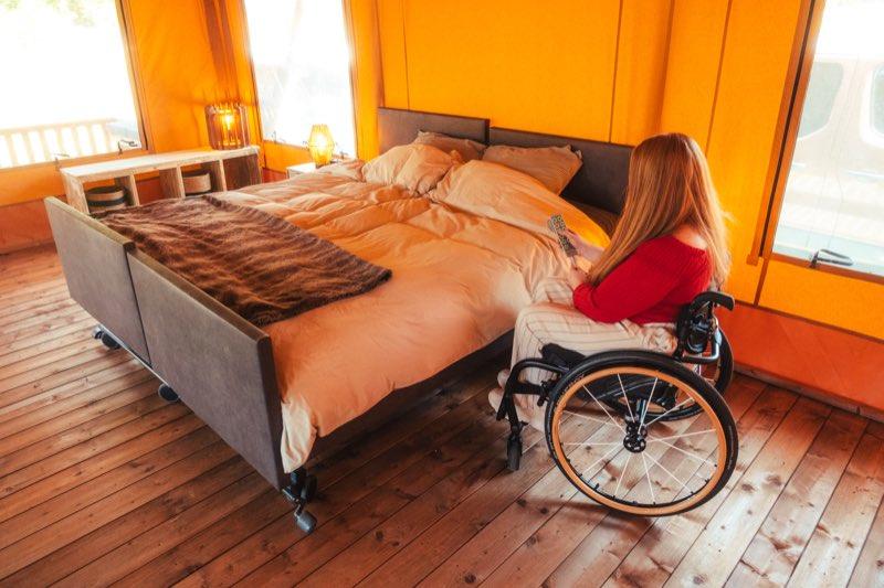 The two single beds pushed together to form a double: a wide bed with sand-coloured linen and a brown throw, standing on castors with a padded foot board. A woman in a manual wheelchair sits beside it holding the bed's handset, its row of buttons used to raise and lower the mattress. Daylight comes through the tent windows behind.