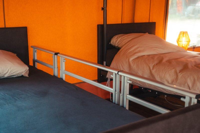 A close view of the removable side rail fitted along the edge of one bed: a light grey aluminium rail with two horizontal bars and upright supports, clamped to the bed frame and standing about a hand's width above the mattress. The second bed is behind it with its duvet turned back.