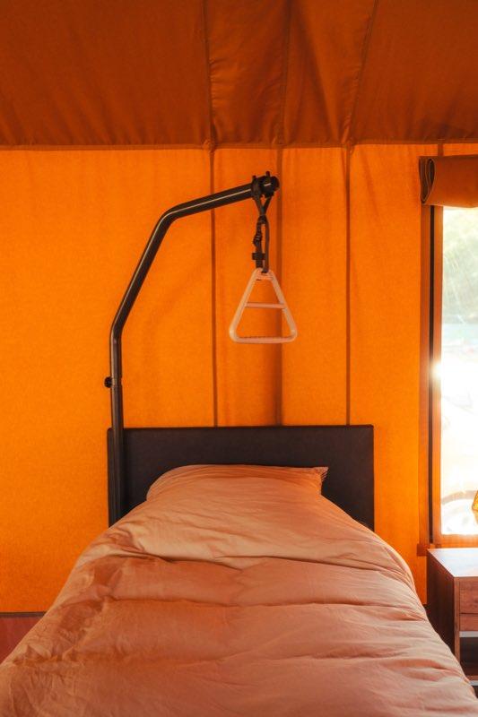 The lifting pole at the head of a bed: a black steel post rises from the floor beside the headboard and curves out over the mattress, with a white triangular trapeze handle hanging from it on an adjustable black strap, within easy reach of someone lying down. The tent's orange canvas wall and a window are behind.
