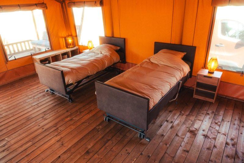 The same two beds set apart as separate singles, each with its own headboard, sand-coloured duvet and upholstered foot board, standing on castors on the wooden floor. The metal high-low frame is visible beneath the nearer bed, and a wide stretch of clear floor runs between and around them for transfers.