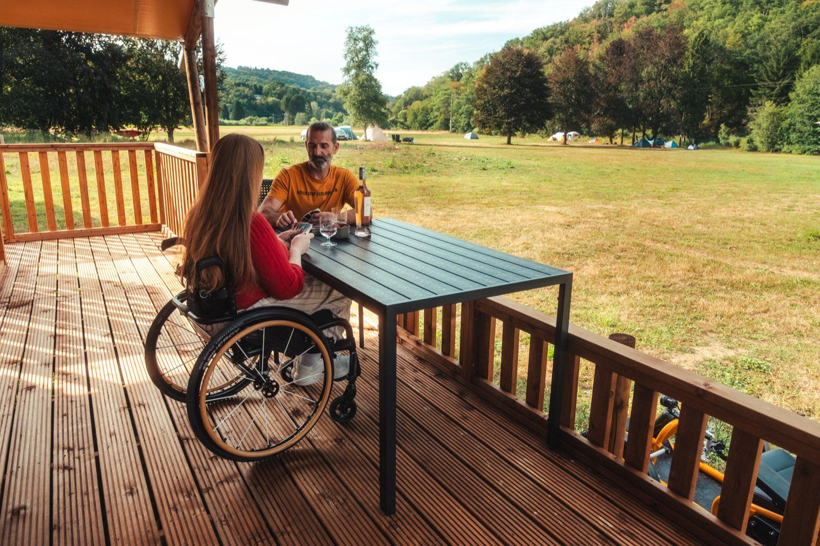 The view from the tent's covered deck: a woman in a manual wheelchair and a man in an orange T-shirt play cards at a dark table, a bottle and two glasses beside them. Beyond the wooden railing a wide mown meadow runs down to the treeline, with small tents pitched along the far edge and the wooded hills of the Ardennes rising behind.