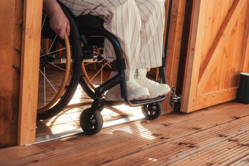Close-up at the tent's doorway: a manual wheelchair with a gold-rimmed wheel rolls over a flat metal threshold strip between the indoor floorboards and the outdoor decking. There is no step or lip — the two floors meet level — and the wide timber door frame and open wooden door stand to the right.