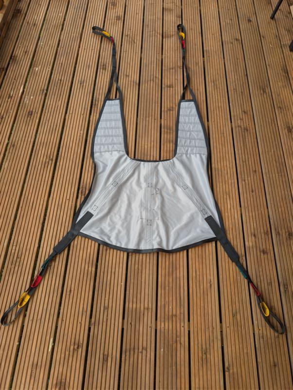 The size L sling laid flat on the deck boards: a grey fabric sling shaped like a vest, with a padded head section, two shoulder straps and two longer leg straps, each ending in colour-coded loops in yellow, red and blue that clip onto the hoist's spreader bar.