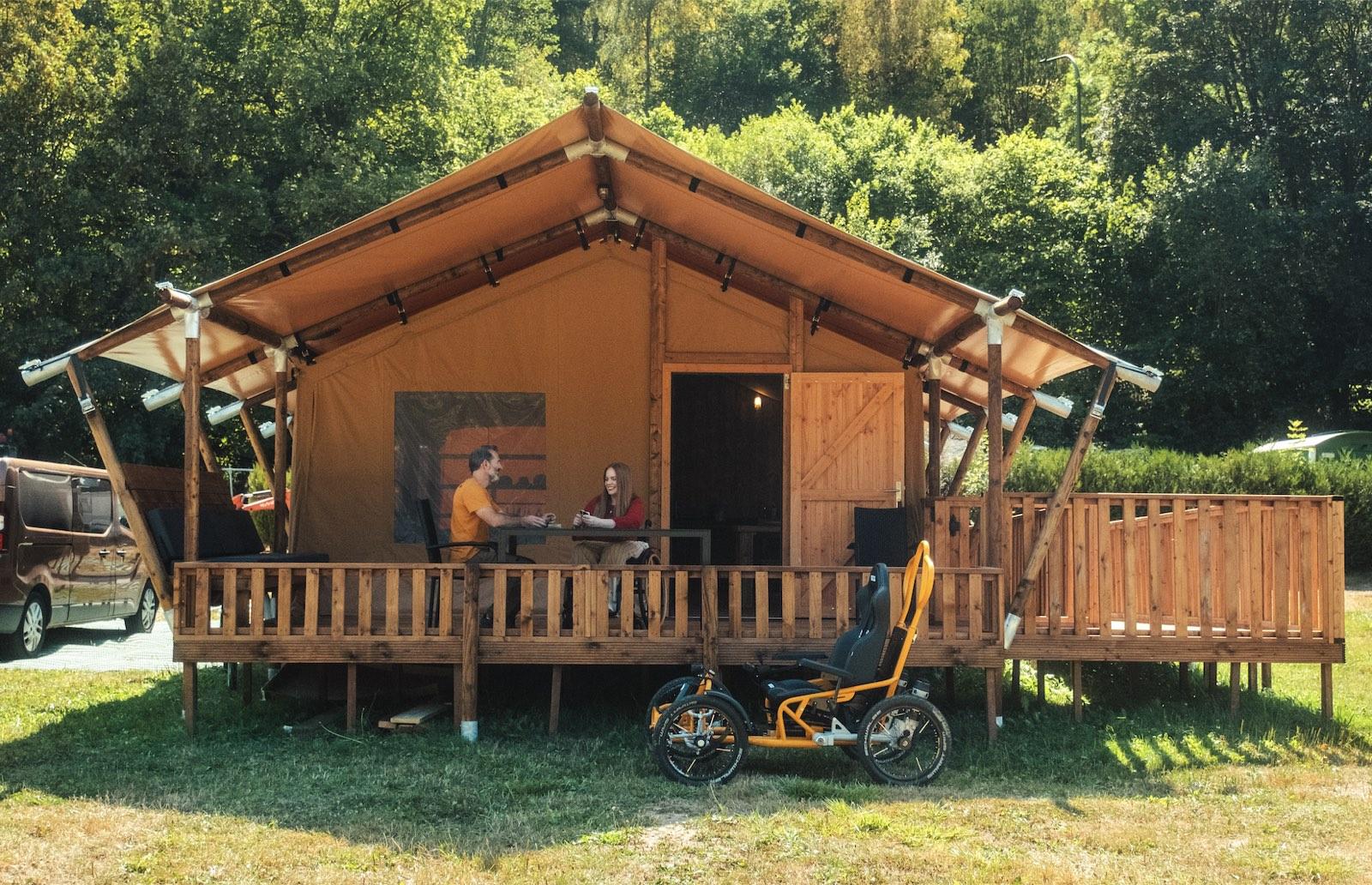 The Piaterra safari tent seen from the front: a canvas roof on a timber frame over a raised wooden deck with a railing. A man in an orange T-shirt and a woman in a wheelchair play cards at a table on the deck, and the orange Quadrix all-terrain wheelchair is parked on the grass in front. A long ramp with a wooden railing runs off to the right, a van is parked on the left, and green woodland rises behind.