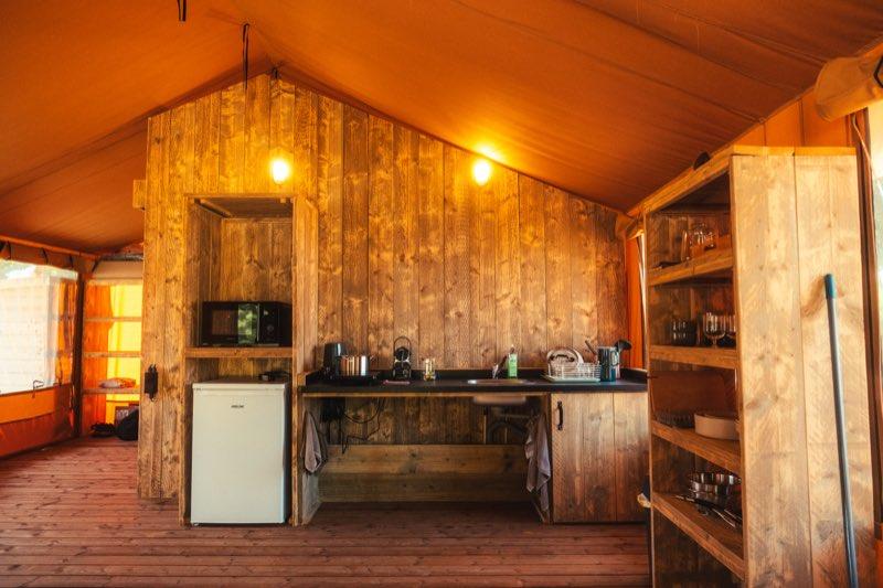 The kitchenette along a reclaimed-timber wall: a dark worktop with an inset sink and hob, the space beneath it left completely open so a wheelchair can roll under. A microwave sits in a recess to the left above a white under-counter fridge, and an open shelving unit with pans, plates and glasses stands to the right.