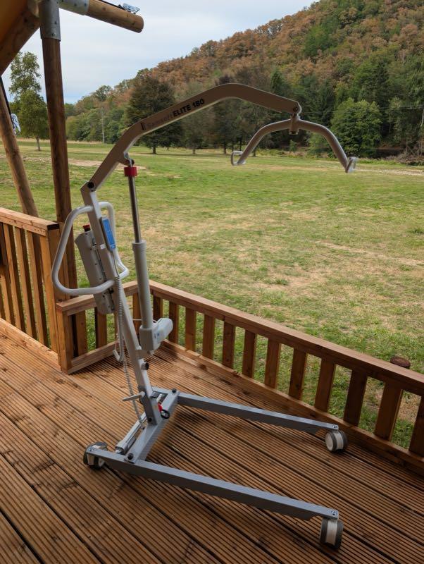 The mobile hoist standing on the deck, its curved spreader bar marked ELITE 180 with a strap hook at each end. A grey mast rises from a wide wheeled base with two splayed legs on castors, and the handset hangs on a coiled cable beside the push handle. The meadow and hillside are behind.