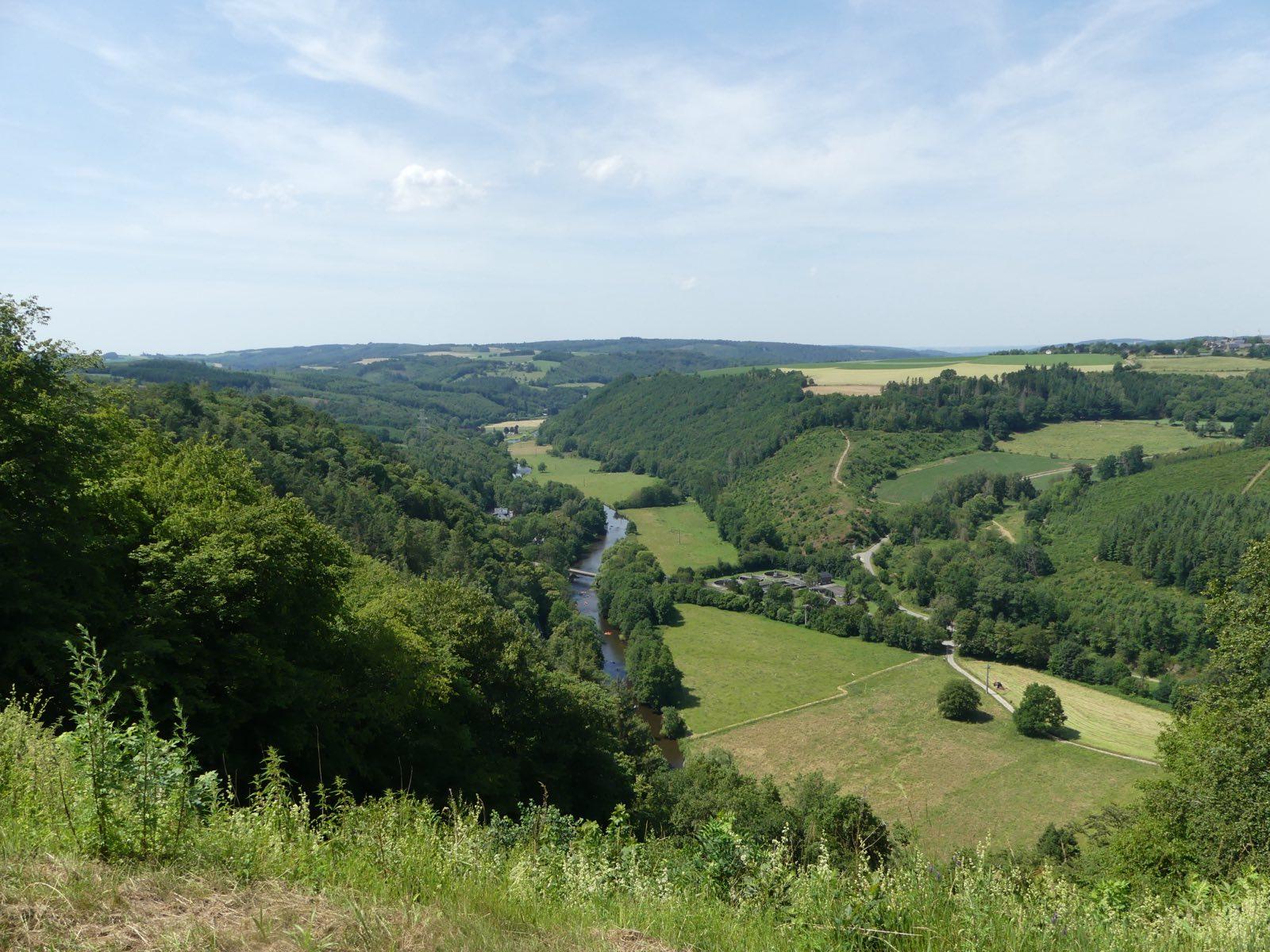 A high viewpoint over the Ourthe valley: the river winds through a deep green valley between steep wooded slopes, with mown meadows, hedgerows and a narrow lane on the valley floor, and rolling forested hills stretching to the horizon under a pale blue summer sky.