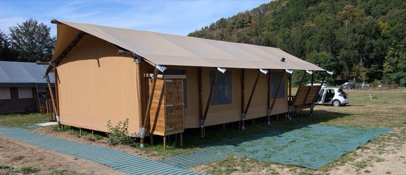 The safari tent seen from the parking area: interlocking green access matting is laid across the grass in a wide path running from the parking space to the tent, which stands on short stilts beneath a broad canvas roof. A white car is parked at the right with its boot open, and wooded hills rise behind the field.