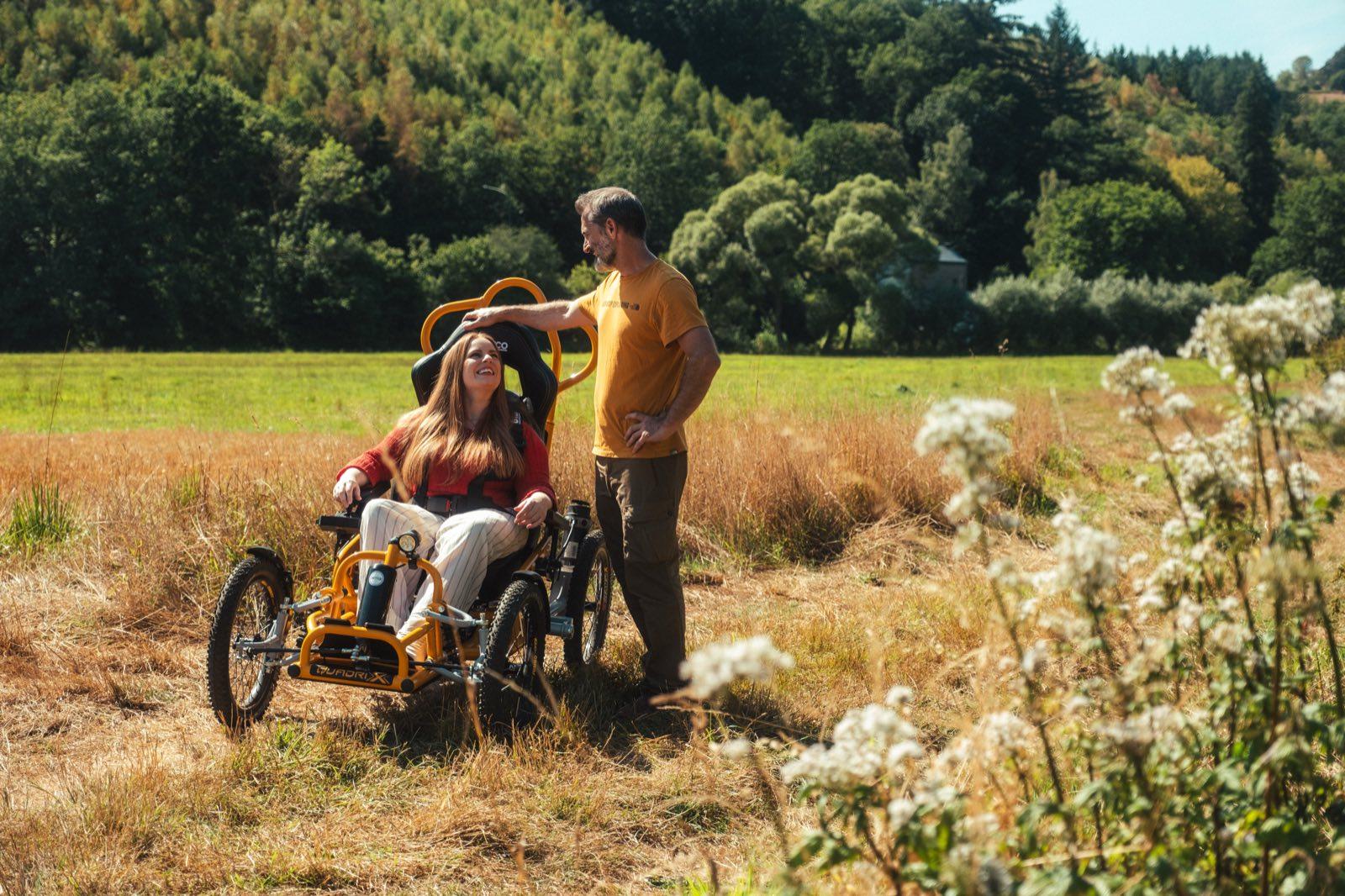 A woman with long red hair drives the orange Quadrix all-terrain wheelchair through a sunlit meadow of tall dry grass, smiling up at a man in an orange T-shirt who walks beside her with one hand resting on the seat's roll bar. The Quadrix runs on four chunky off-road tyres; wooded hills and white wildflowers frame the field behind them.