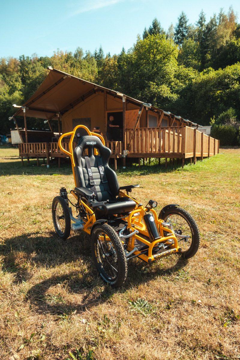 The orange Quadrix all-terrain wheelchair parked on the grass in front of the Piaterra safari tent, seen close up: four knobbly off-road tyres, a black bucket seat and a tubular roll bar. Behind it the tent's canvas roof, wooden deck and railing, with forest rising beyond.
