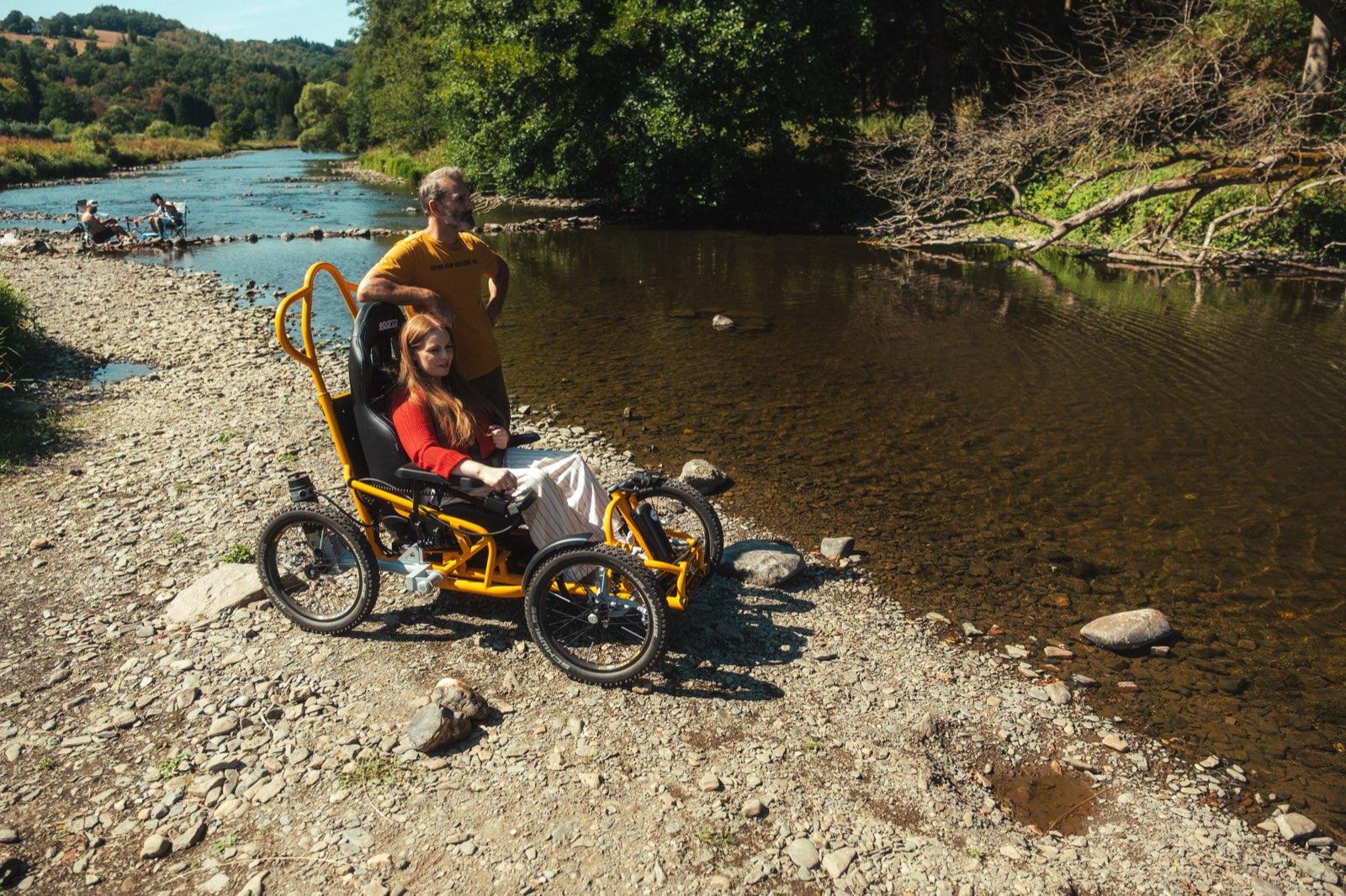 The orange Quadrix all-terrain wheelchair standing on the stony shore of the river Ourthe, its rider seated with a blanket over her legs and a man standing beside her, both looking out across the shallow water to the wooded bank opposite.