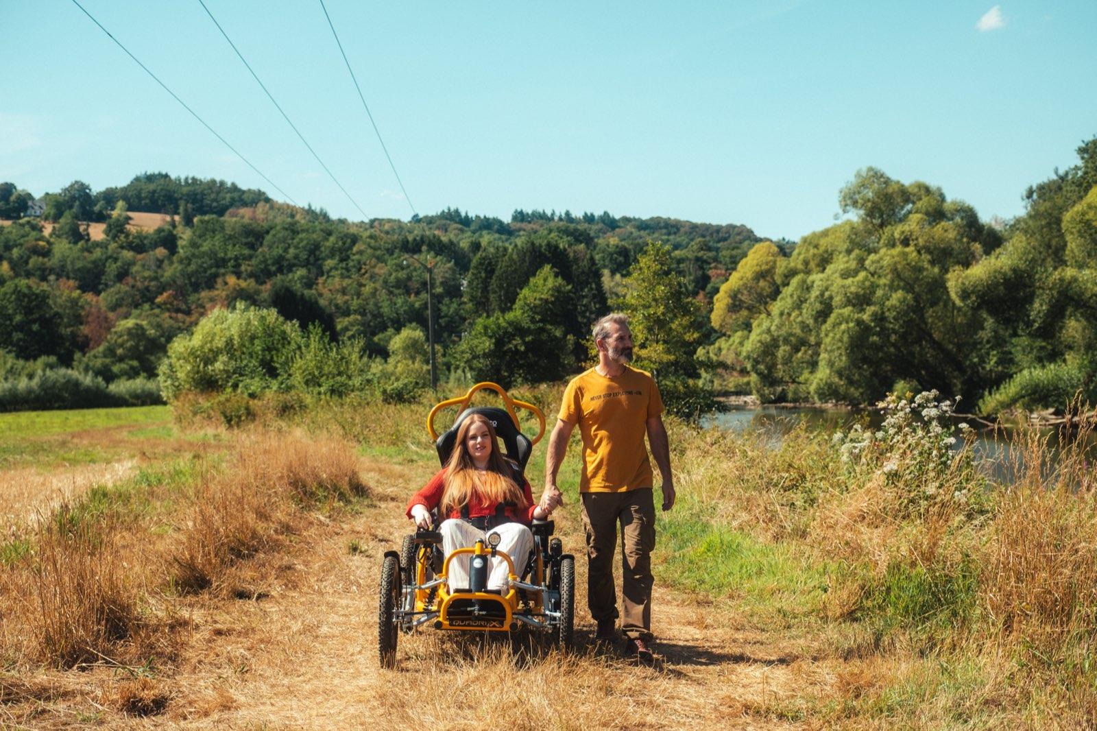 A woman with long red hair drives the orange Quadrix all-terrain wheelchair along a dry earth track through a sunlit meadow, holding hands with a man in an orange T-shirt walking beside her. The chair runs on four chunky off-road tyres, with the river Ourthe, willow trees and the wooded hills of the Ardennes behind them.