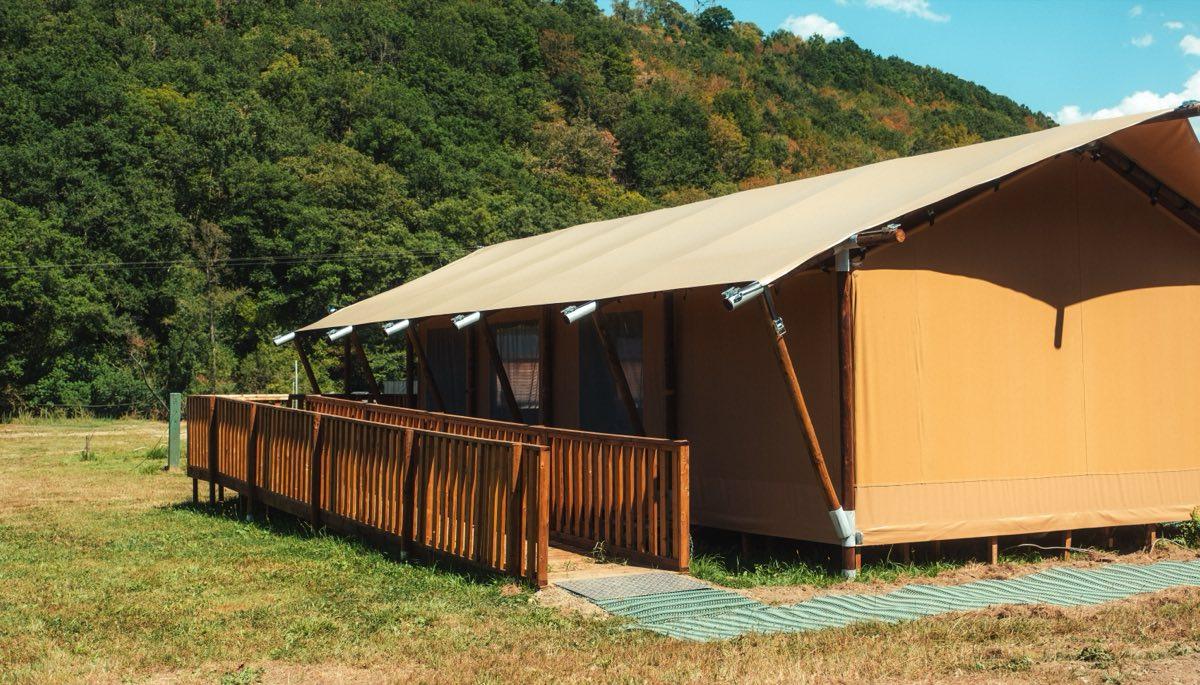 A long wooden ramp with a handrail on both sides rises gently from the grass to the tent&rsquo;s raised deck, turning once along the way. Green access matting covers the ground at the foot of the ramp, and the tent&rsquo;s canvas roof and wooden posts fill the right of the frame with wooded hills behind.