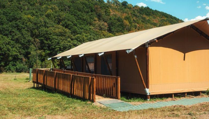 A long wooden ramp with a handrail on both sides rises gently from the grass to the tent's raised deck, turning once along the way. Green access matting covers the ground at the foot of the ramp, and the tent's canvas roof and wooden posts fill the right of the frame with wooded hills behind.
