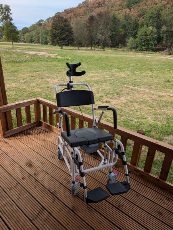 The mobile shower and toilet chair standing on the wooden deck: a white aluminium frame on four braked castors, with a padded black seat with a commode opening, a black backrest, padded flip-up armrests, height-adjustable black footplates and a curved black headrest on an adjustable post. Green meadow and a wooded hillside lie behind.