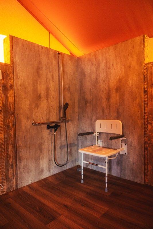The step-free shower corner: a chrome riser rail with a handheld shower head and a black mixer tap on a concrete-look wall, with a horizontal chrome grab bar beside it. A white folding shower seat with a backrest and padded armrests is mounted on the adjoining wall on height-adjustable legs, and a round drain sits flush in the dark wood floor.