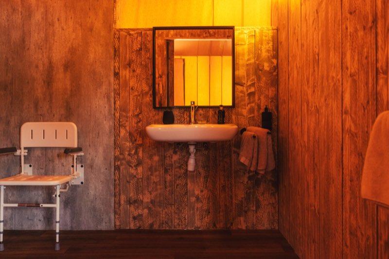 A wall-hung sink with the space underneath left open so a wheelchair can roll up to it, the exposed trap set back against the timber wall. A square mirror hangs above, with a tap, soap and a glass on the narrow rim, folded towels on a rail to the right, and the white folding shower seat visible on the wall at the left.
