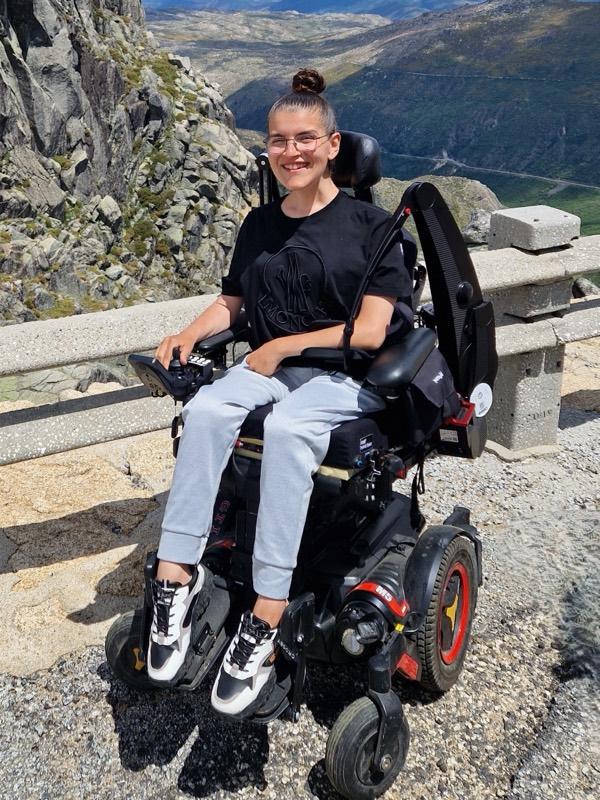 Asmae smiling in her power wheelchair at a mountain viewpoint, with a wide valley behind her.