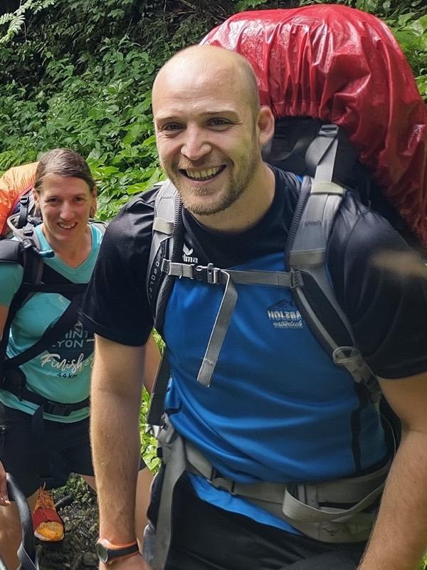 Seb smiling on a green forest trail, carrying a large hiking pack, with fellow walkers behind him.