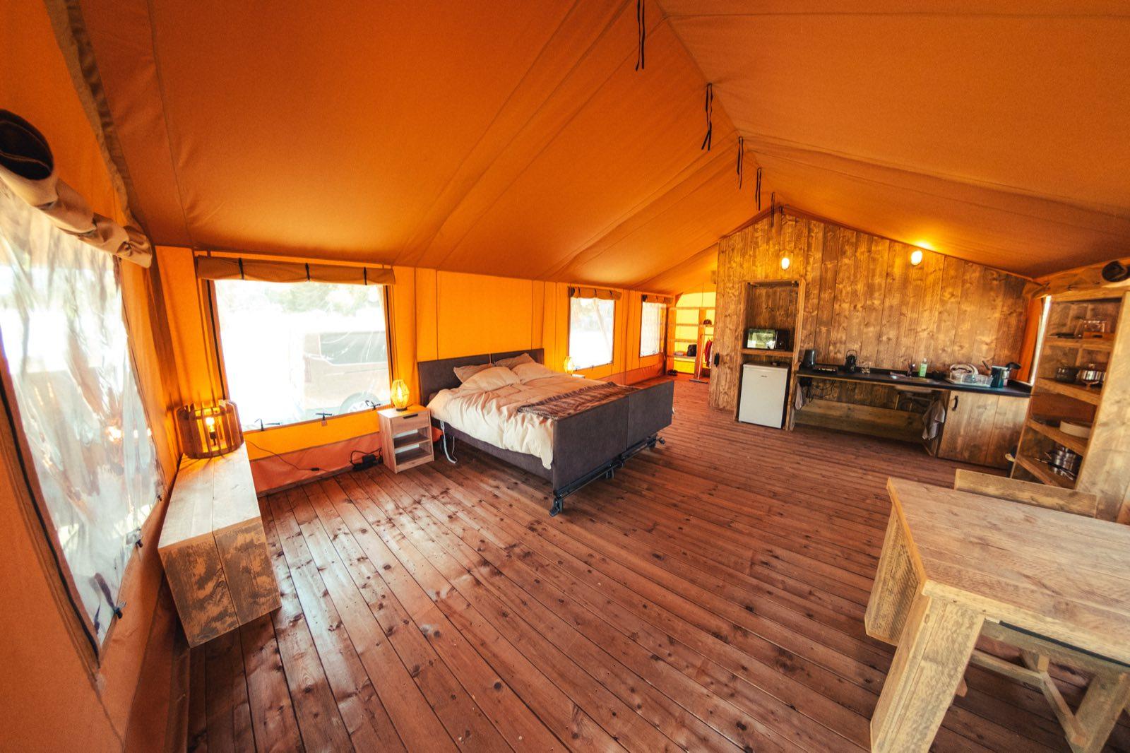 Inside the safari tent: a bright open room under a high canvas roof with a wooden plank floor. An electric profiling bed on a metal frame stands in the middle with clear floor space around it and underneath. Along the right-hand wall a wooden kitchenette with worktop, sink, small fridge and microwave; a dining table in the foreground and large windows letting in daylight.