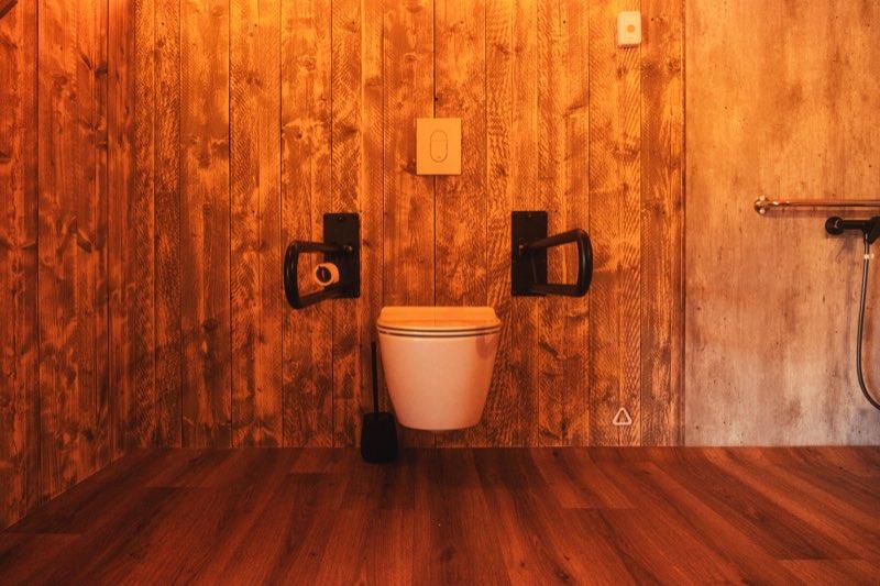 A wall-hung toilet with a wooden seat, mounted on a reclaimed-timber wall with a black folding grab bar on each side at matching height, both lowered into the horizontal position. The flush plate is set into the wall above, a toilet brush stands to the left, and the dark wood floor beneath is clear, with open space to the right for a side transfer.